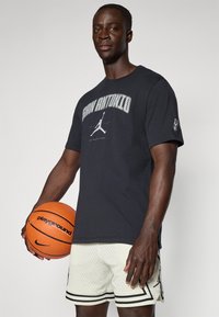 Juodas medvilninis TATARAS su "San Antonio" grafika ir Jumpman logotipu; derinamas su baltais perforuotais šortais su juodais akcentais. Laiko oranžinį krepšinį.