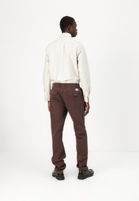 Pantalons en coton marron avec une coupe décontractée, dotés de deux poches arrière et d'un logo en patch, associés à une chemise à manches longues blanche à rayures.