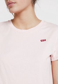 Jasnoróżowa bawełniana koszulka z krótkimi rękawami, z małym czerwonym logo Levi's po lewej stronie na klatce piersiowej. Gładka faktura, klasyczny krój z okrągłym dekoltem.