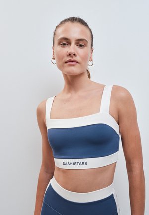 Junge Frau mit zurückgebundenen Haaren trägt ein blau-weißes Sportoberteil und hoch taillierte Leggings mit DASH- und STARS-Logo oben.