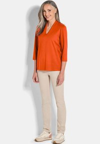 Rotes V-Ausschnitt-Oberteil mit Dreiviertelärmeln, kombiniert mit beigen Slim-Fit-Hosen und hellen Schuhen. Weicher Stoff mit glatter Textur.