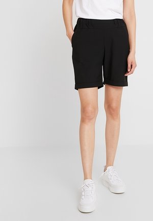 Osoba w czarnych szortach z podwiniętym dołem, białej koszulce włożonej w spodenki i białych sznurowanych sneakersach stoi na jednolitym tle.