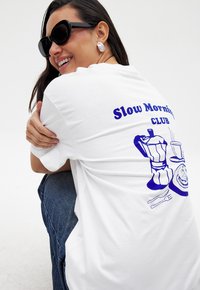 T-shirt en coton blanc avec un imprimé graphique bleu d'une cafetière et d'articles de petit-déjeuner au dos, associé à un jean en denim foncé.