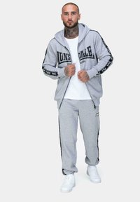 Šedá mikina na zips s čiernym logom, spárovaná s nohavicami na cvičenie v rovnakom štýle. Obsahuje pruhované akcenty po stranách a biele tenisky.