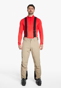 Beige Skihosen mit schwarzen Trägern, zwei vorderen Knöpfen, strukturiertem Stoff und schwarzen Akzenten an den Knöcheln. Getragen mit einem roten Langarmoberteil.