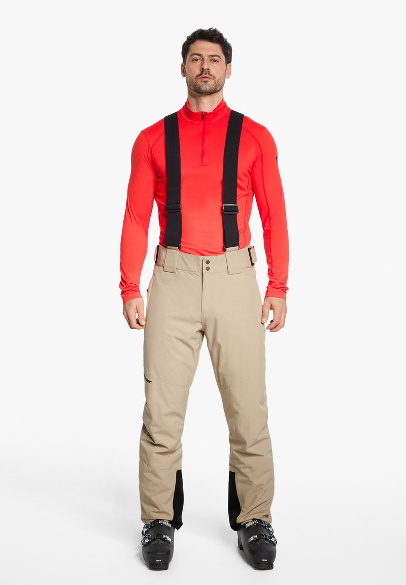 Beige Skihosen mit schwarzen Trägern, zwei vorderen Knöpfen, strukturiertem Stoff und schwarzen Akzenten an den Knöcheln. Getragen mit einem roten Langarmoberteil.