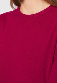 Fuchsia stickad tröja med klassisk rund halsringning, mjuk textur och ribbad detalj runt kragen och ärmarna.