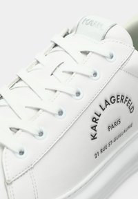 Sapatilha de pele branca com bico redondo, atacadores planos, e logótipo em relevo: "KARL LAGERFELD" e detalhes de endereço em preto. Acabamento suave.