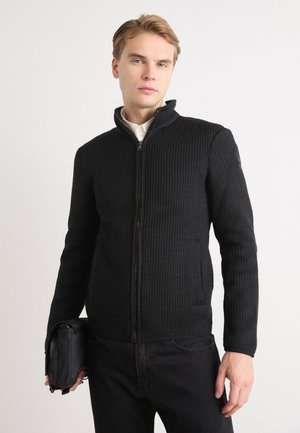 Giacca nera con zip, caratterizzata da un motivo testurizzato a coste. Presenta un colletto alto, tasche laterali e una zip frontale liscia. Indossata con pantaloni neri.