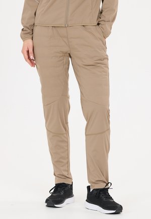 Personne portant un pantalon et une veste de sport beige, les mains dans les poches, associée à des chaussures de course noires sur un fond uni clair.