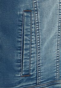 Džínová bunda v světlé modré barvě, s obdélníkovou kapsou s prošívanými okraji a viditelnou texturou, doplněná kontrastním hnědým nitěm.