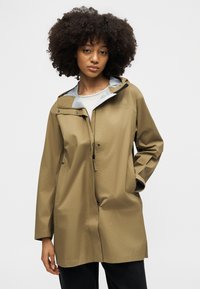 Donna con capelli ricci che indossa una giacca impermeabile color sabbia con cappuccio, chiusure con zip e bottoni a pressione, in piedi con una mano in tasca.