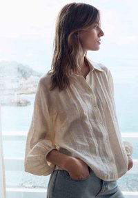 Frau mit langen braunen Haaren in einem locker sitzenden beigen Hemd und Jeans schaut zur Seite und steht drinnen an einem Fenster mit Meerblick.