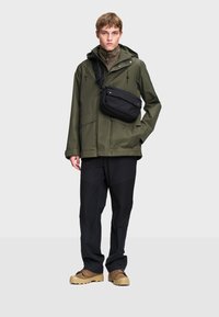 Zelená vodoodolná bunda s veľkými vreckami, skombinovaná s čiernymi nohavicami s textúrovaným dizajnom a hnedými topánkami. Malá čierna crossbody taška.