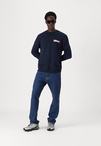 Marineblå genser med rund hals og logo-patch, kombinert med blå denimjeans og grå joggesko. Modell med svarte solbriller.