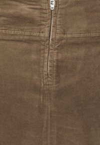 Gros plan d'une fermeture éclair métallique argentée partiellement dézippée sur un tissu en daim marron avec des détails de couture visibles.