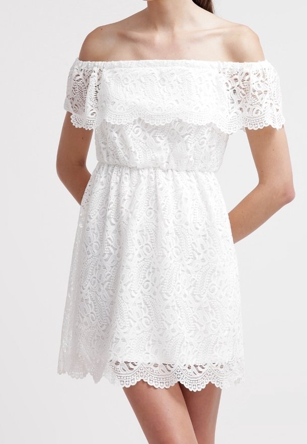 Femme portant une robe blanche en dentelle épaules dénudées avec des bords festonnés et une taille élastique, debout devant un fond uni.