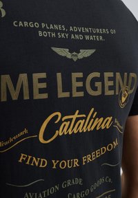 Crna pamučna majica s zlatnim tekstom i grafikom krila. Tekst uključuje "JA LEGEND", "Catalina" i "PRONAĐI SVOJU SLOBODU." Vintage dizajn.