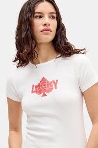 Biela bavlnená tričko s poškodeným červeným logom "LUCKY" a dizajnom srdca na prednej strane. Krátke rukávy a priliehavý strih.