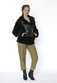 Zwarte jas van nepbont met lederen accenten, ritssluiting en grote kraag, gecombineerd met geel-zwart geruite broek en zwarte enkellaarzen.