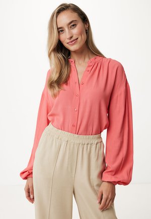 Blusa di corallo leggera con maniche lunghe a palloncino, chiusura con bottoni e scollatura leggermente arricciata, abbinata a pantaloni beige con vita elastica.