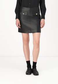 Schwarzer Leder-Minirock mit Fronttaschen, goldenen Knopfakzenten und einer taillierten Passform. Getragen mit schwarzen Socken und Slippern.