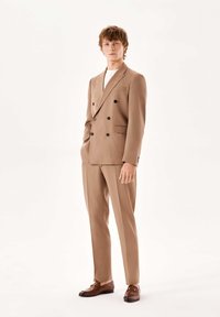 Traje beige de doble botonadura con botones negros, pantalones ajustados y mocasines marrones. La camiseta blanca complementa el diseño minimalista.
