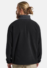 Giacca di pile nera con colletto alto, tessuto strutturato e vestibilità ampia. Presenta una cucitura sul retro e maniche lunghe con polsini.