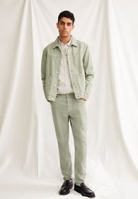 Ljusgrön denimjacka med tryckknappar, matchande ljusgröna byxor och svarta loafers. Modellen står mot en texturerad vit bakgrund.