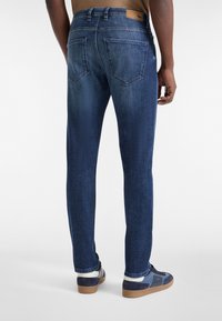 Jeans de mezclilla azul oscuro con un corte ajustado, que cuentan con una cinturilla estándar y dos bolsillos traseros. La tela muestra una sutil textura y un ligero desgastado.
