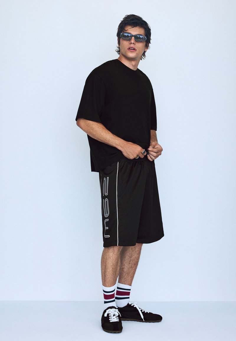 Jeune homme portant des lunettes de soleil noires, un t-shirt noir oversize, un short noir avec des lettres blanches, des chaussettes rayées et des baskets noires, debout contre un mur clair.