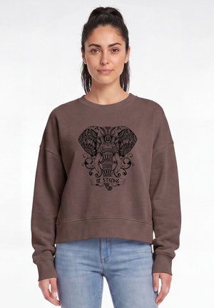 Frau mit dunklen Haaren im Pferdeschwanz trägt braunen Sweatshirt mit aufwändigem Elefantendesign und "Be Strong" Text, kombiniert mit hellblauen Jeans.