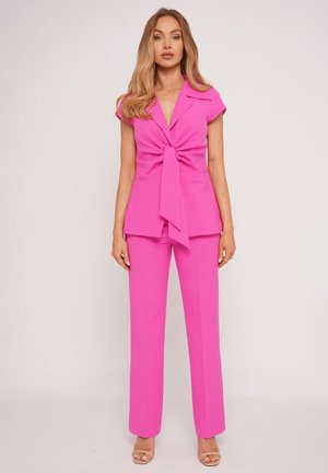 Kobieta o długich blond włosach, ubrana w jaskrawy różowy bezrękawnik z wiązaniem z przodu oraz pasujące szerokie spodnie, stojąca na jednolitym tle.