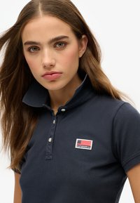 Marine polo skjorte lavet af tekstureret stof med krave. Har et lille flagpatch med "SUPERDRY" label på venstre bryst.