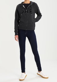 Felpa con zip grigia scura con logo "GAP" sul davanti, abbinata a jeans skinny blu scuro e sneakers bianche con dettagli marroni.
