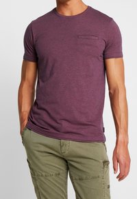 Camiseta de manga corta y cuello redondo en tejido burdeos texturizado con un bolsillo en el pecho, combinada con pantalones cargo verde claro.