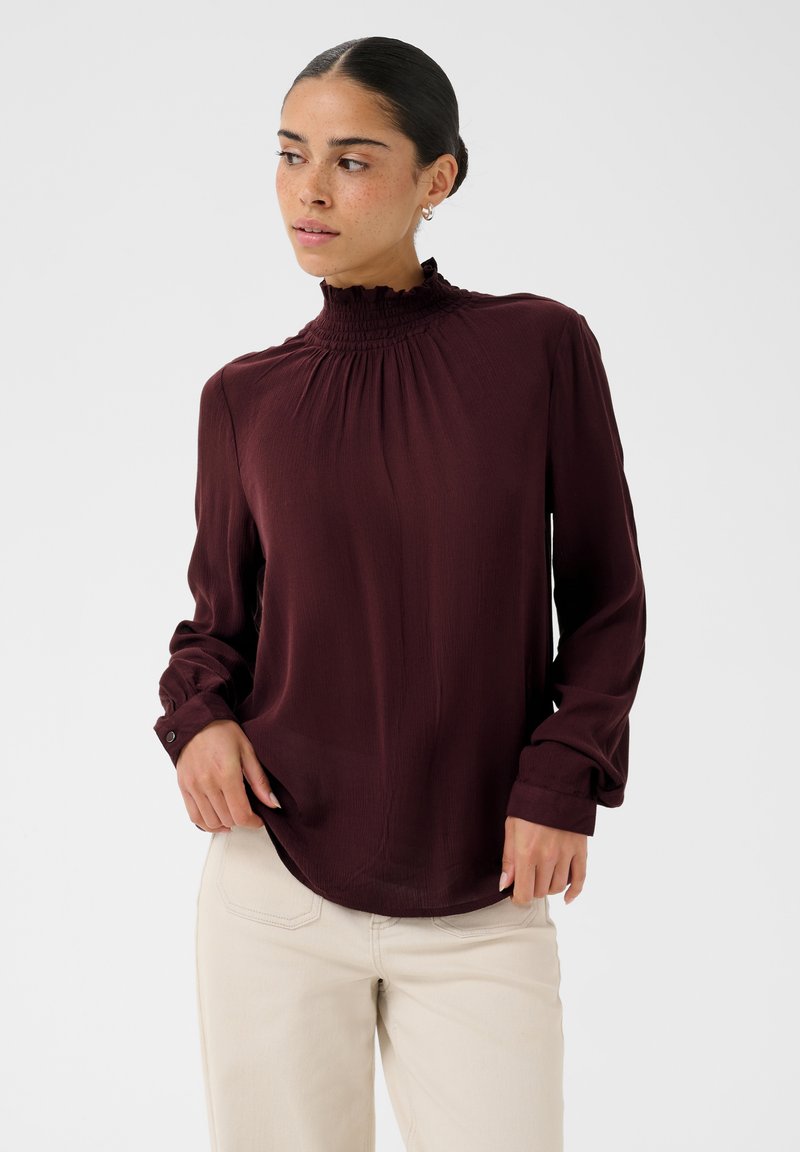 Femme portant un chemisier violet foncé à manches longues avec un col haut à volants et un pantalon blanc cassé sur un fond clair uni.