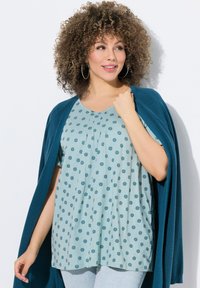 Světle modrý top s květinovým vzorem, zdobený plisováním. Tyrkysový cardigan přes ramena. Modelka má kudrnaté vlasy.