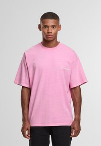 Nooruke mehine lühikeste juustega, seljas lahtine roosa T-särk ja mustad püksid, seismas ühetoalise heledashalliga taustal.