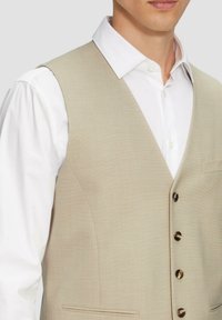 Beige Weste mit glatter Textur, scharfem V-Ausschnitt-Design, fünf dunklen Knöpfen und einer Tasche. Über einem weißen Hemd mit Knopfleiste getragen.