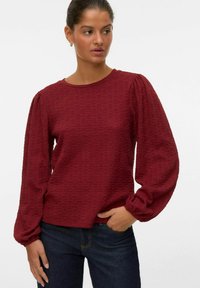 Bordeauxfärgad, texturerad långärmad topp med puffärmar och rund halsringning, kombinerad med mörka jeans. Diskret mönster i hela tyget.