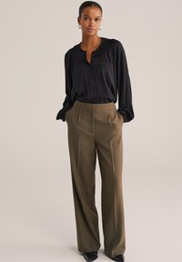 Egy nő áll, kezét zsebébe dugva, fekete hosszúujjú blúzt, olíva zöld széles szárú nadrágot, fekete hegyes cipőt és ezüst fülbevalót viselve.