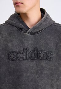 Juodas dėvėtas džemperis su kišene kengūrai ir siuvinėtu "adidas" logotipu. Pagamintas iš tekstūruoto audinio ir turi reguliuojamą raištelį ant kapučino.