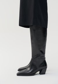 Stivali neri in pelle al ginocchio con punta affusolata e tacco quadrato, caratterizzati da una texture liscia e un design minimalista.