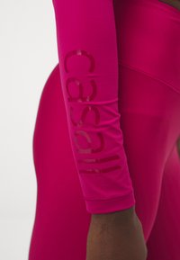 Prilagođena atletska majica dugih rukava od svijetloružičaste tkanine. Sadrži sjajni "casall" logo otisnut duž rukava. Glatka tekstura, rastezljiv materijal.