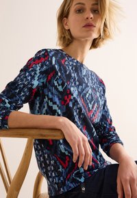 Marineblauwe lange mouw top met geometrisch patroon in blauw en rood. Geribbelde halslijn en manchetten, met drie zwarte accentknopen aan de onderrand.