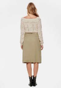 Pull en tricot beige avec un motif ajouré, associé à une jupe verte olive arrivant au genou et des bottines noires, vu de dos.