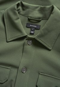 Chemise verte avec un col structuré, dotée d'une fermeture à boutons, de deux poches et d'une texture de tissu lisse. Étiquette de taille L visible.
