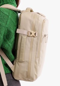 Beige batoh s polstrovanými popruhy, bočním zipovým kapsou a texturovanými materiály, nošený na zeleném svetru.