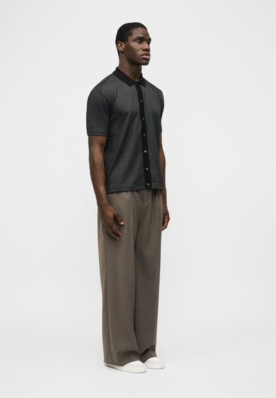 Άνδρας που στέκεται φορώντας κοντομάνικο μαύρο πουκάμισο με σχέδια και κουμπιά, φαρδιά καφέ παντελόνια και λευκά αθλητικά παπούτσια, με φόντο απλή ανοιχτή επιφάνεια.